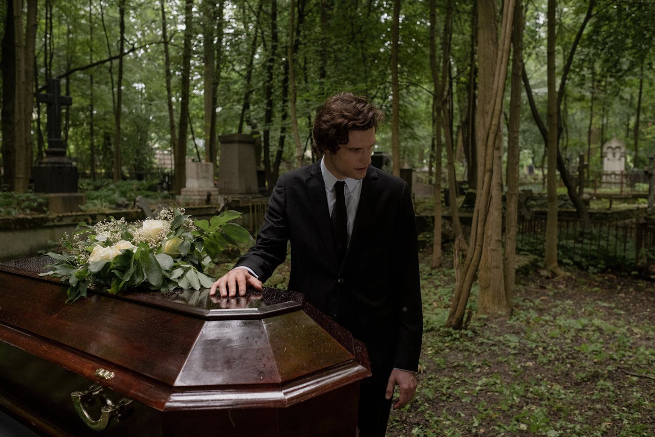 A man in a black suit mourns at a funeral in a lush green cemetery, leaning on the coffin.
